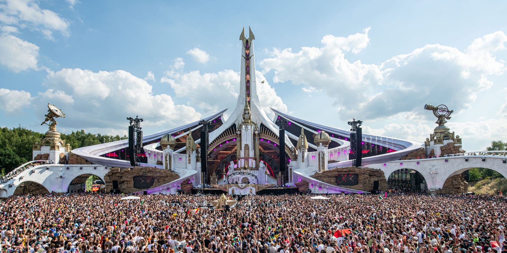 Tomorrowland's ornate fantasy mainstage in Boom, Belgium, with pyrotechnic rockets launching into the golden evening sky above a massive crowd.