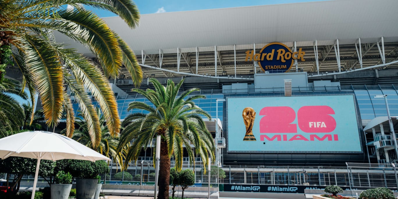 Hard Rock Stadium in Miami displaying a FIFA World Cup 2026 branded billboard, framed by palm trees on a sunny day.