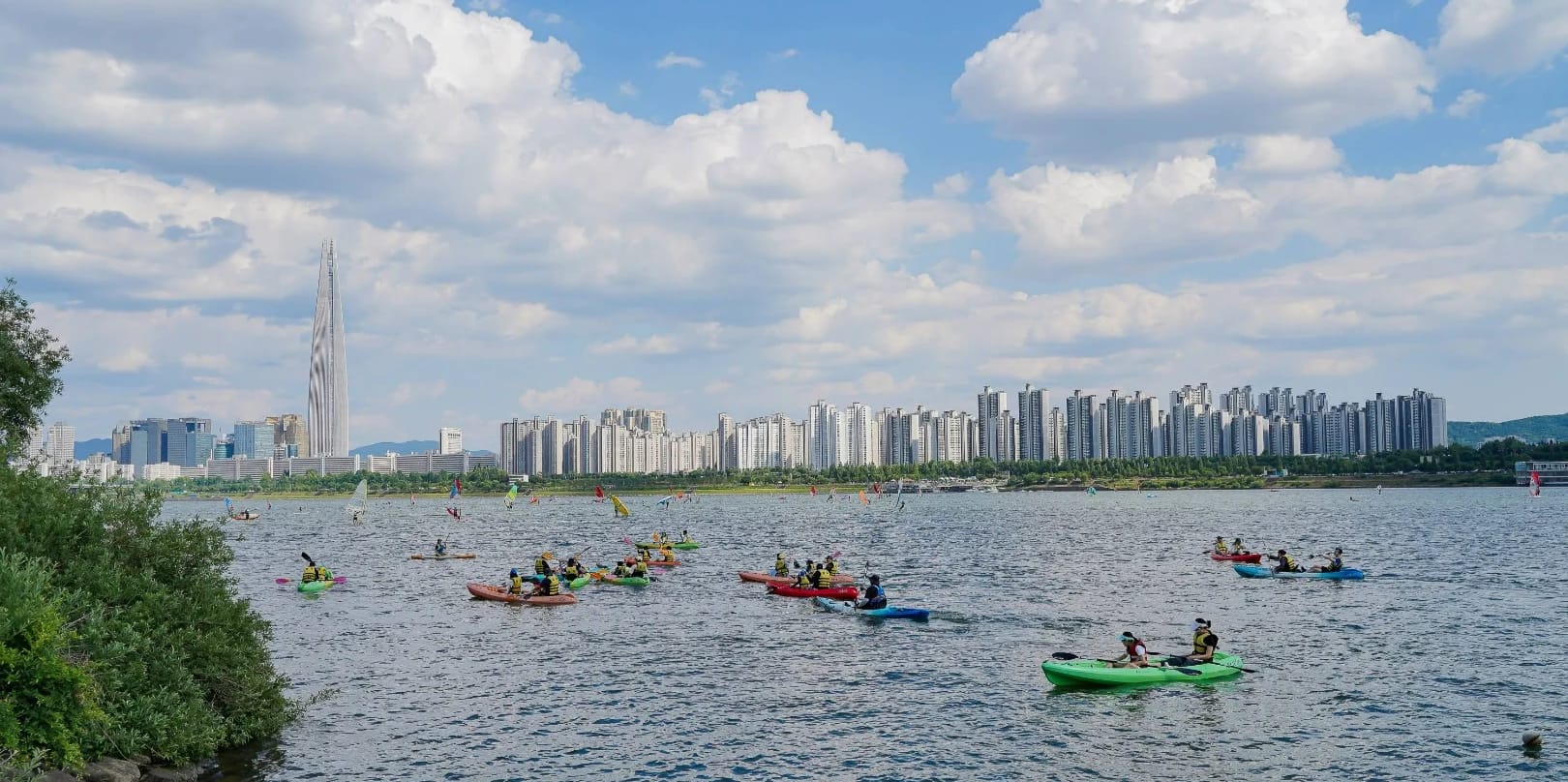 Kayakers paddling on the Han River with Lotte World Tower and Seoul's skyline in the background on a sunny day.